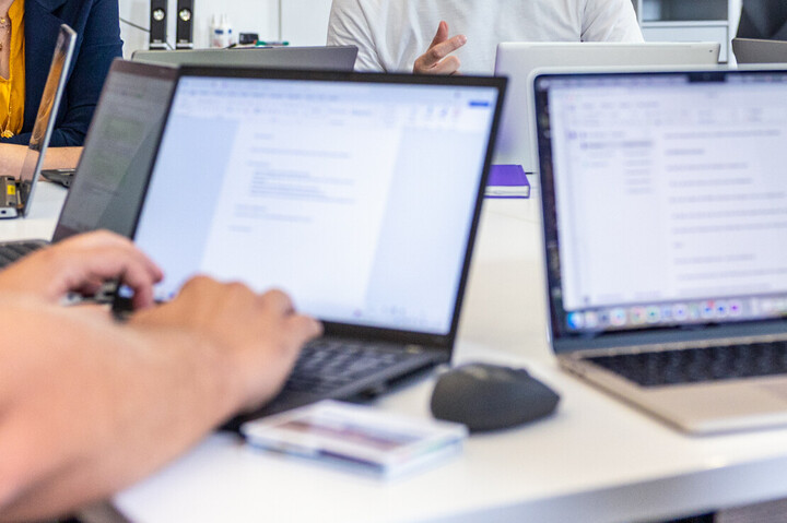 Mitarbeitende sitzen mit ihren Laptops in einer Besprechung. Im Fokus ist ein Mitarbeiter, der spricht.