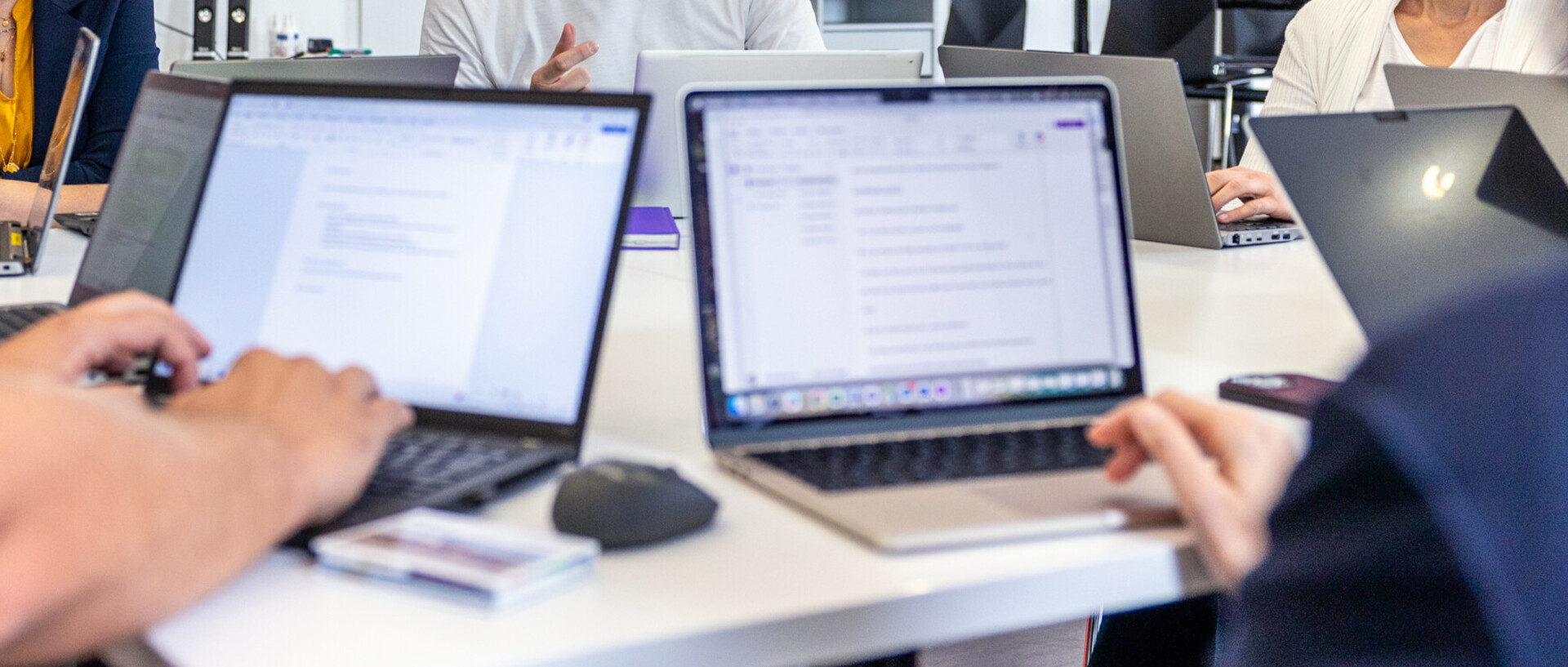 Mitarbeitende sitzen mit ihren Laptops in einer Besprechung. Im Fokus ist ein Mitarbeiter, der spricht.