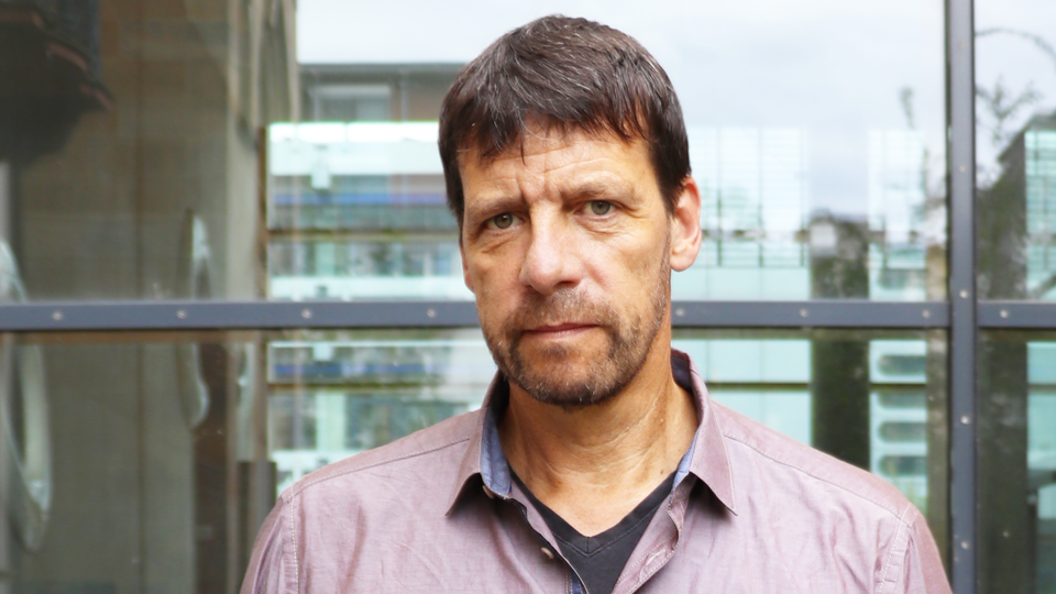 Porträt von Professor Krause, der vor einer Glaswand steht, ein lilafarbenes Hemd trägt und in die Kamera schaut.