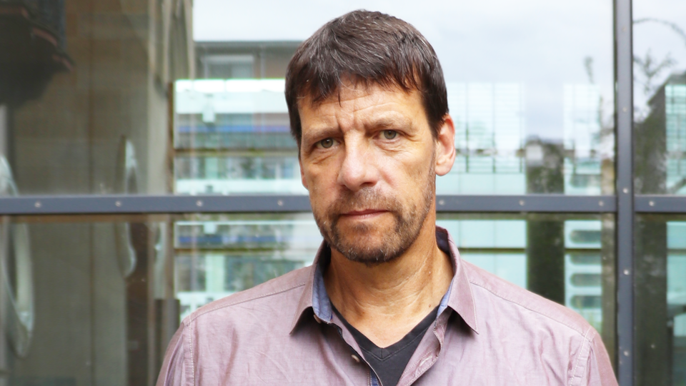 Porträt von Professor Krause, der vor einer Glaswand steht, ein lilafarbenes Hemd trägt und in die Kamera schaut.