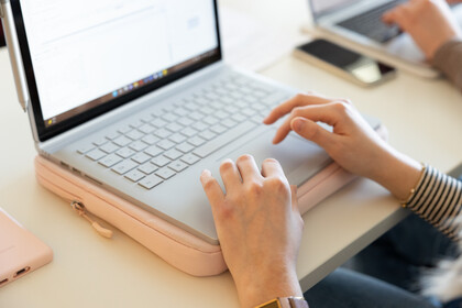Aufgeklappter Laptop und eine Person, die darauf schreibt, vermutlich eine Frau. Man sieht nur die Hände.