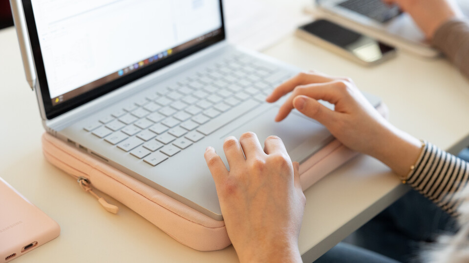 Aufgeklappter Laptop und eine Person, die darauf schreibt, vermutlich eine Frau. Man sieht nur die Hände.