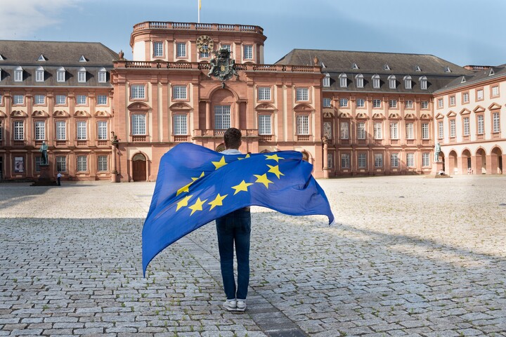 Eine Person steht mit dem Rücken zur Kamera und schau Richtung Mannheimer Schloss. Auf ihrem Rücken eine wehende Europa-Flagge.