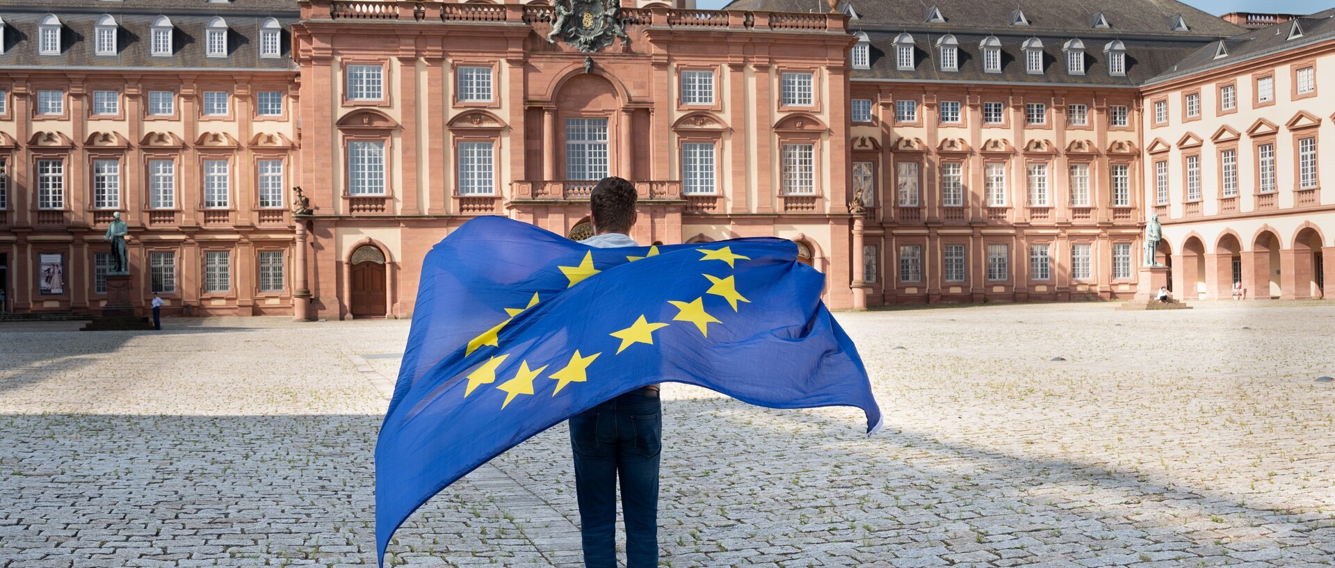 Eine Person steht mit dem Rücken zur Kamera und schau Richtung Mannheimer Schloss. Auf ihrem Rücken eine wehende Europa-Flagge.