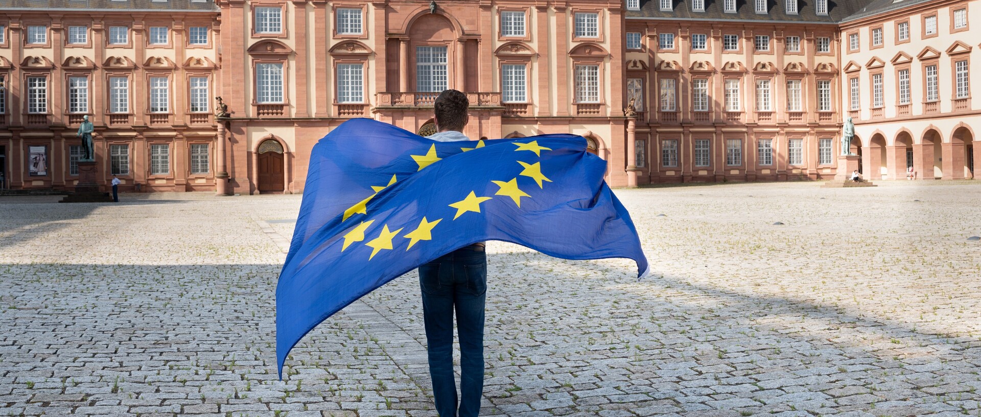 Eine Person steht mit dem Rücken zur Kamera und schau Richtung Mannheimer Schloss. Auf ihrem Rücken eine wehende Europa-Flagge.