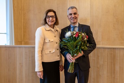 Zwei lächelnde Personen in schicker Kleidung stehen nebeneinander vor einer braunen Wand. Die Person rechts, Thomas Fetzer, hält einen Blumenstrauß in der Hand.