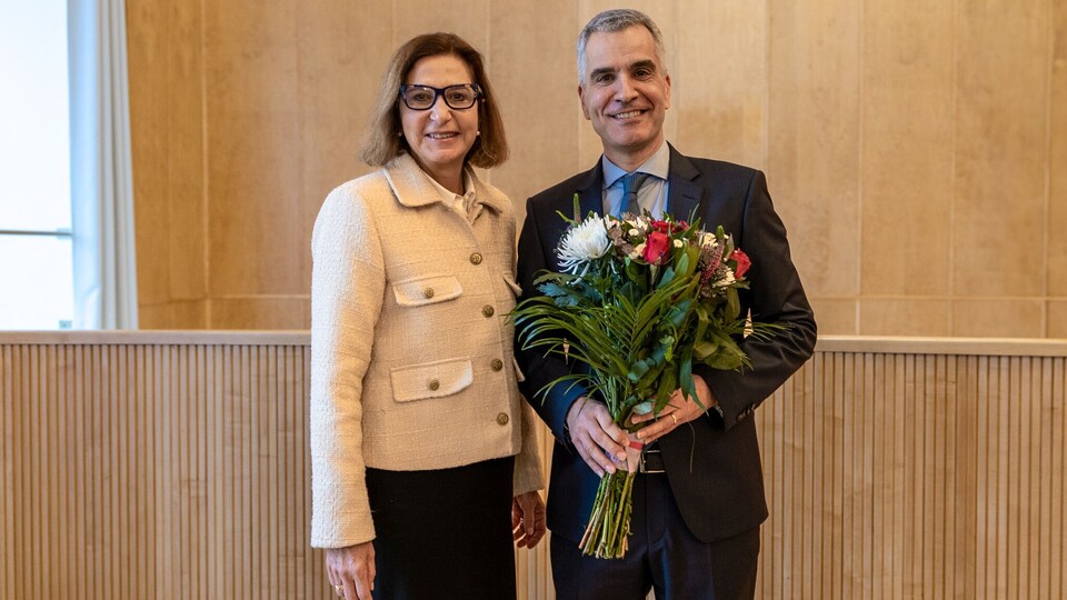Zwei lächelnde Personen in schicker Kleidung stehen nebeneinander vor einer braunen Wand. Die Person rechts, Thomas Fetzer, hält einen Blumenstrauß in der Hand.