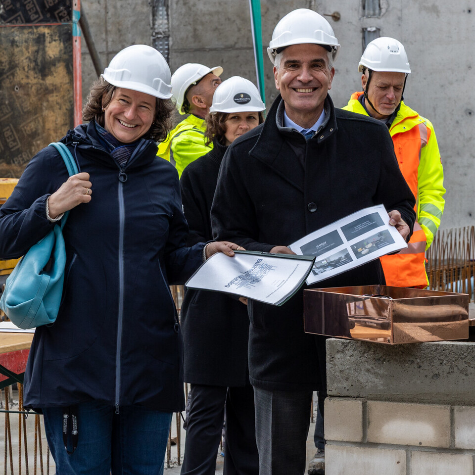 Grundsteinlegung auf einer Baustelle: Katrin Schoppa und Thomas Fetzer mit weißen Schutzhelmen halten Dokumente und stehen lächelnd vor einer Kupferkapsel. Im Hintergrund Bauarbeiter in Warnwesten.