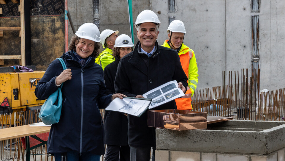 Grundsteinlegung auf einer Baustelle: Katrin Schoppa und Thomas Fetzer mit weißen Schutzhelmen halten Dokumente und stehen lächelnd vor einer Kupferkapsel. Im Hintergrund Bauarbeiter in Warnwesten.