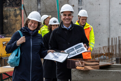Grundsteinlegung auf einer Baustelle: Katrin Schoppa und Thomas Fetzer mit weißen Schutzhelmen halten Dokumente und stehen lächelnd vor einer Kupferkapsel. Im Hintergrund Bauarbeiter in Warnwesten.