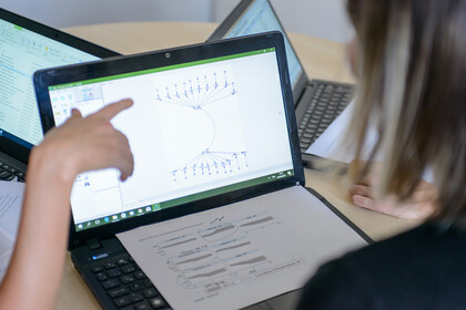 Ein aufgeklappter Laptop steht auf einem Tisch. Eine Person zeigt mit ihrem Finger auf die Grafik, die auf dem Laptop angezeigt wird.