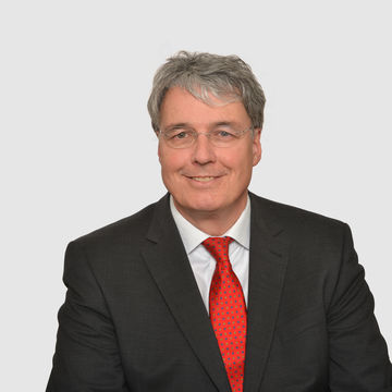 Prof. Heinzl hat graue kurze Haare und eine Brille. Er trägt ein dunkles Jacket, ein weißes Hemd und eine rote Krawatte.