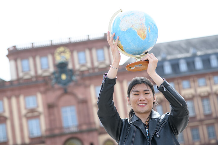 Ein Studierender Hält einen Globus in die Höhe. Im Hintergrund sieht man unscharf das Schloss.