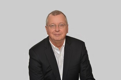 Portrait von Martin Weber. Er trägt ein weißes Hemd sowie einen schwarzen Blazer und sitzt lächelnd vor einer grauen Wand.
