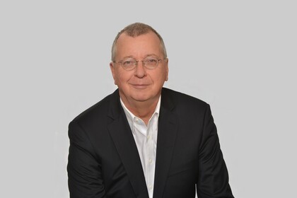 Portrait von Martin Weber. Er trägt ein weißes Hemd sowie einen schwarzen Blazer und sitzt lächelnd vor einer grauen Wand.