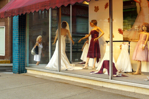 Links überquert ein Mann die Straße. Rechts befindet sich das Schaufenster eines Brautmodegeschäfts. Durch die Ecke des Glases ist eine ältere Dame zu sehen, die ein ausgestelltes Kleid betrachtet.