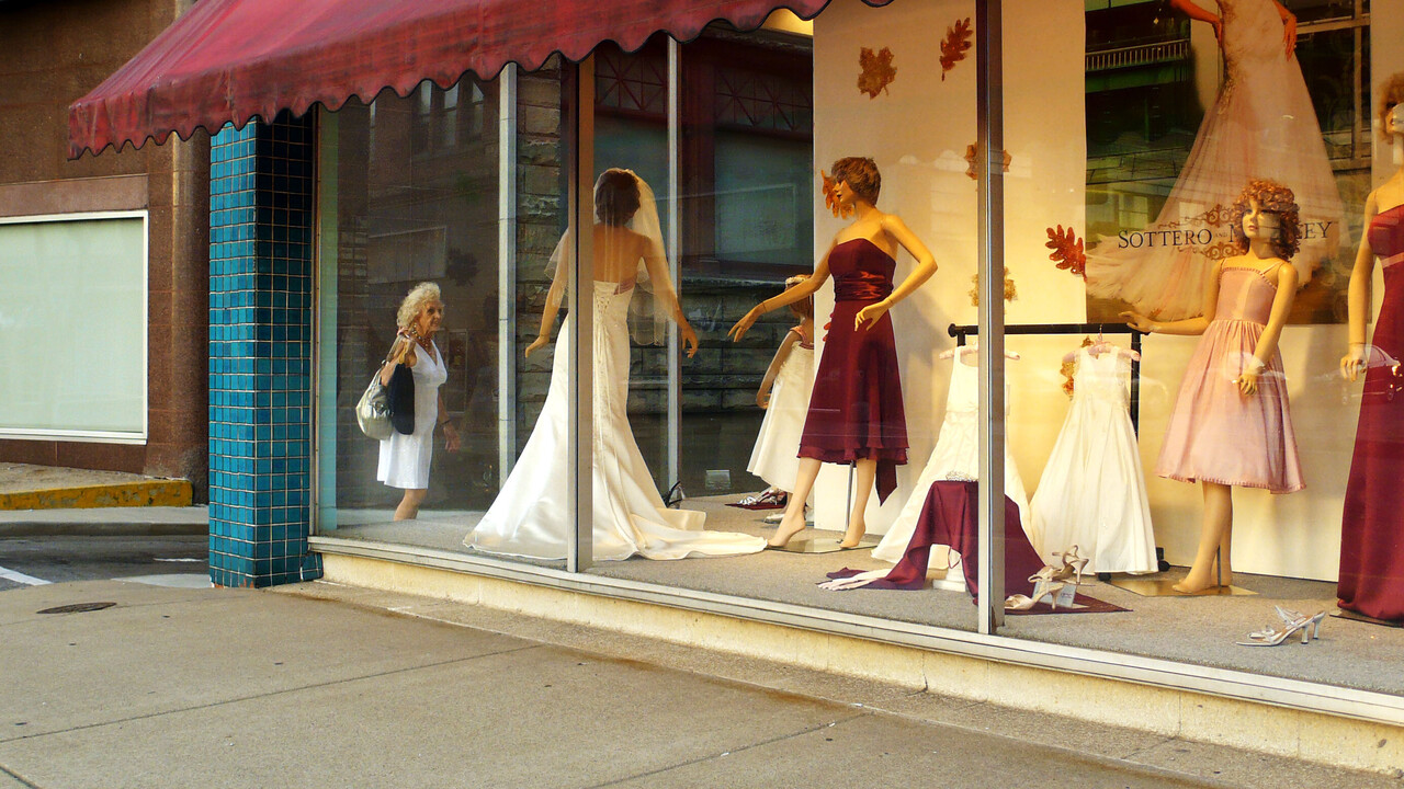 Links überquert ein Mann die Straße. Rechts befindet sich das Schaufenster eines Brautmodegeschäfts. Durch die Ecke des Glases ist eine ältere Dame zu sehen, die ein ausgestelltes Kleid betrachtet.