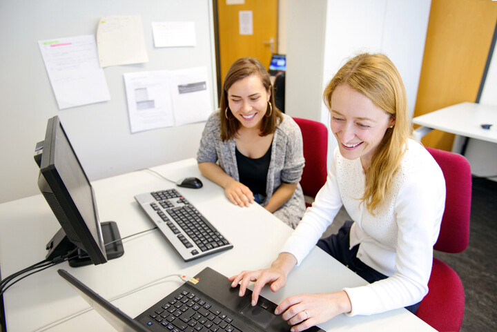 Zwei Frauen sitzen lachend vor einem Laptop mit Bildschirm.