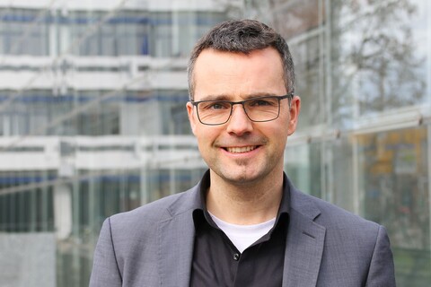 Portrait von Marc Debus. Er trägt ein schwarzes Hemd sowie einen grauen Blazer und steht vor dem A5-Gebäude.