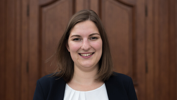 Hannah Rajski. She is wearing a black jacket over a white blouse and is standing in front of a brown door.