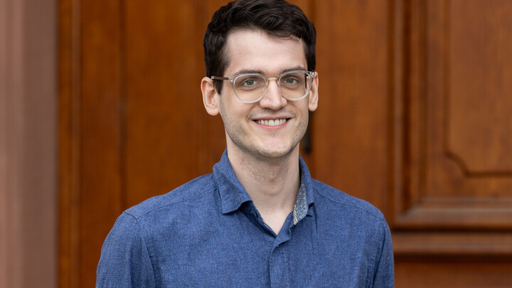 He is wearing a dark blue, jeans-like shirt and is standing in front of an entrance to Mannheim Palace.