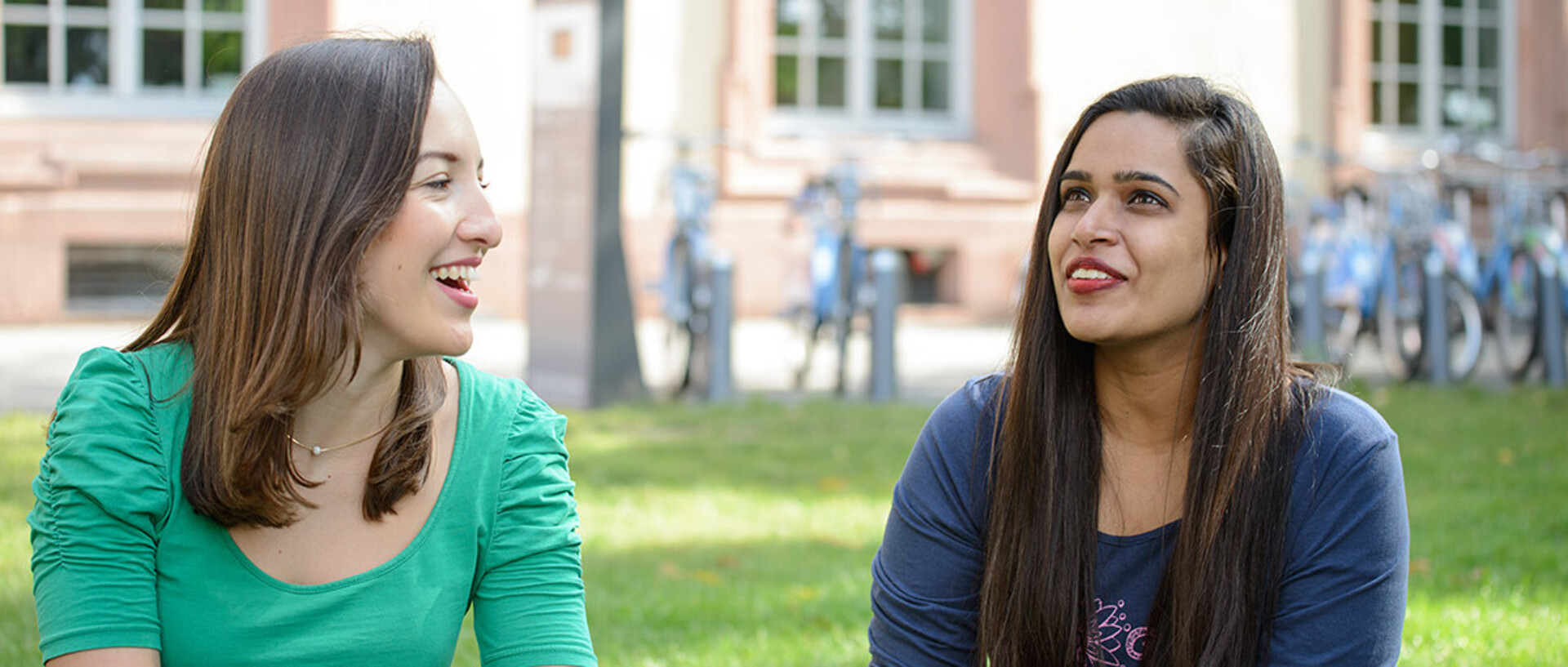 Zwei Studentinnen sitzen auf dem Campus auf der Wiese und unterhalten sich.