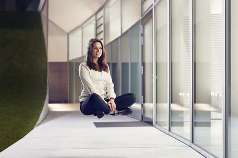 Lächelnde Frau mit braunen schulterlangen Haaren, weißer Bluse und schwarzer Jeans sitzt in Schneidersitz vor einem Haus schwebend in der Luft.
