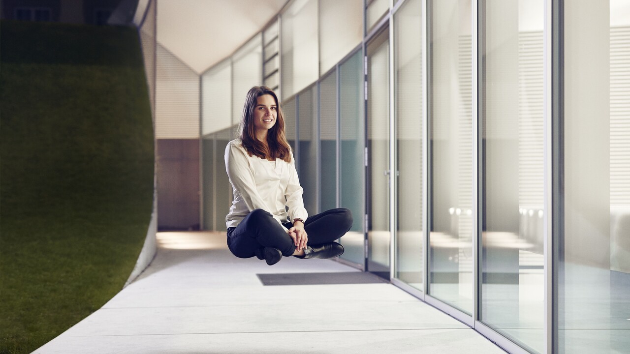 Lächelnde Frau mit braunen schulterlangen Haaren, weißer Bluse und schwarzer Jeans sitzt in Schneidersitz vor einem Haus schwebend in der Luft.