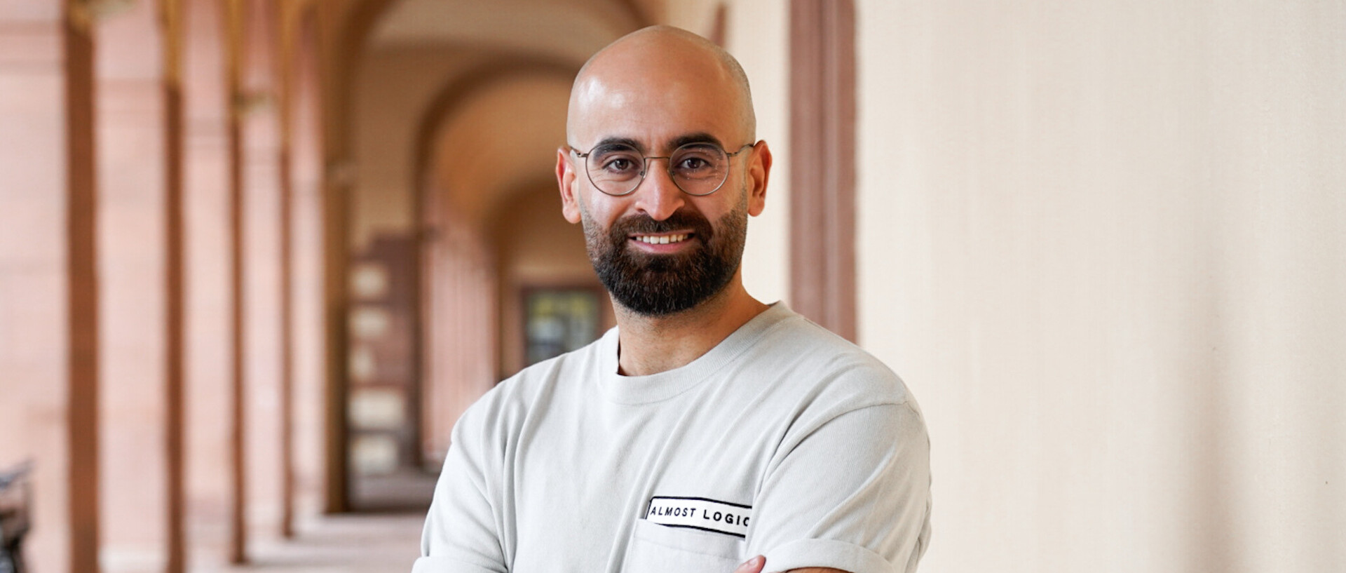 Hassan Abdolaziz stands smiling in an entrance hall of the Schloss. He wears a beige T-shirt and crosses his arms in front of his chest.