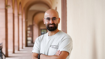 Hassan Abdolaziz stands smiling in an entrance hall of the Schloss. He wears a beige T-shirt and crosses his arms in front of his chest.