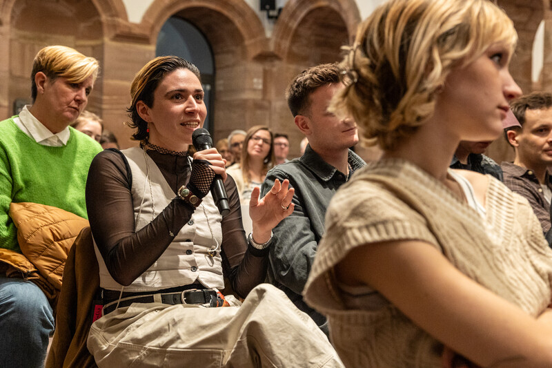 Zuschauerin spricht während der Hauptveranstaltung der Diversity Week in ein Mikrofon und stell eine Frage. Sie gestikuliert dabei mit den Händen. Im Hintergrund sind zudem andere Zuschauer zu sehen.