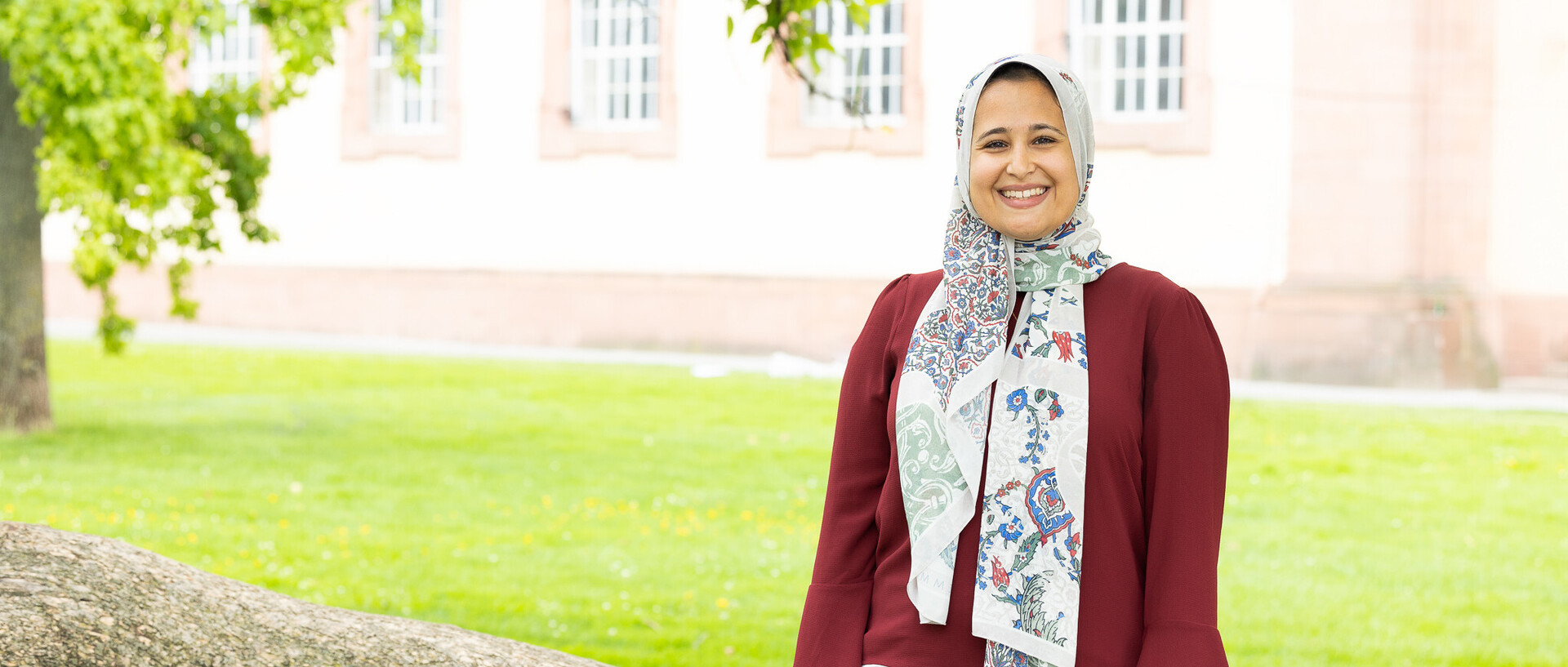 Alaa Elfawal stands smiling in front of a tree. She is wearing a colorful headscarf, a red blouse, and white trousers. The Schloss is in the background.
