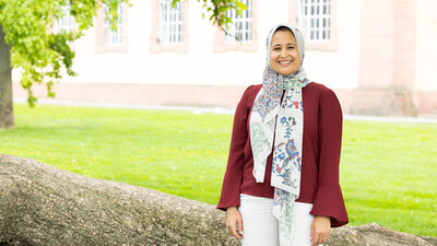 Alaa Elfawal stands smiling in front of a tree. She is wearing a colorful headscarf, a red blouse, and white trousers. The Schloss is in the background.