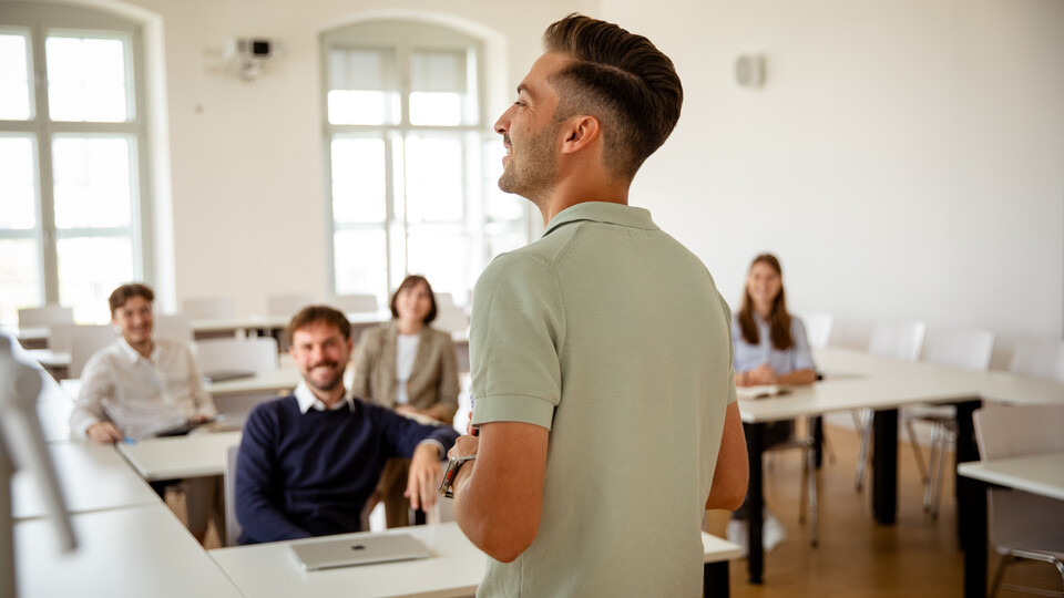 Eine Person in einem Poloshirt steht vorne in einem Semianrraum und redet. Fünf weitere Personen sitzen im Raum und sehen die Person an.