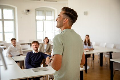 Eine Person in einem Poloshirt steht vorne in einem Semianrraum und redet. Fünf weitere Personen sitzen im Raum und sehen die Person an.