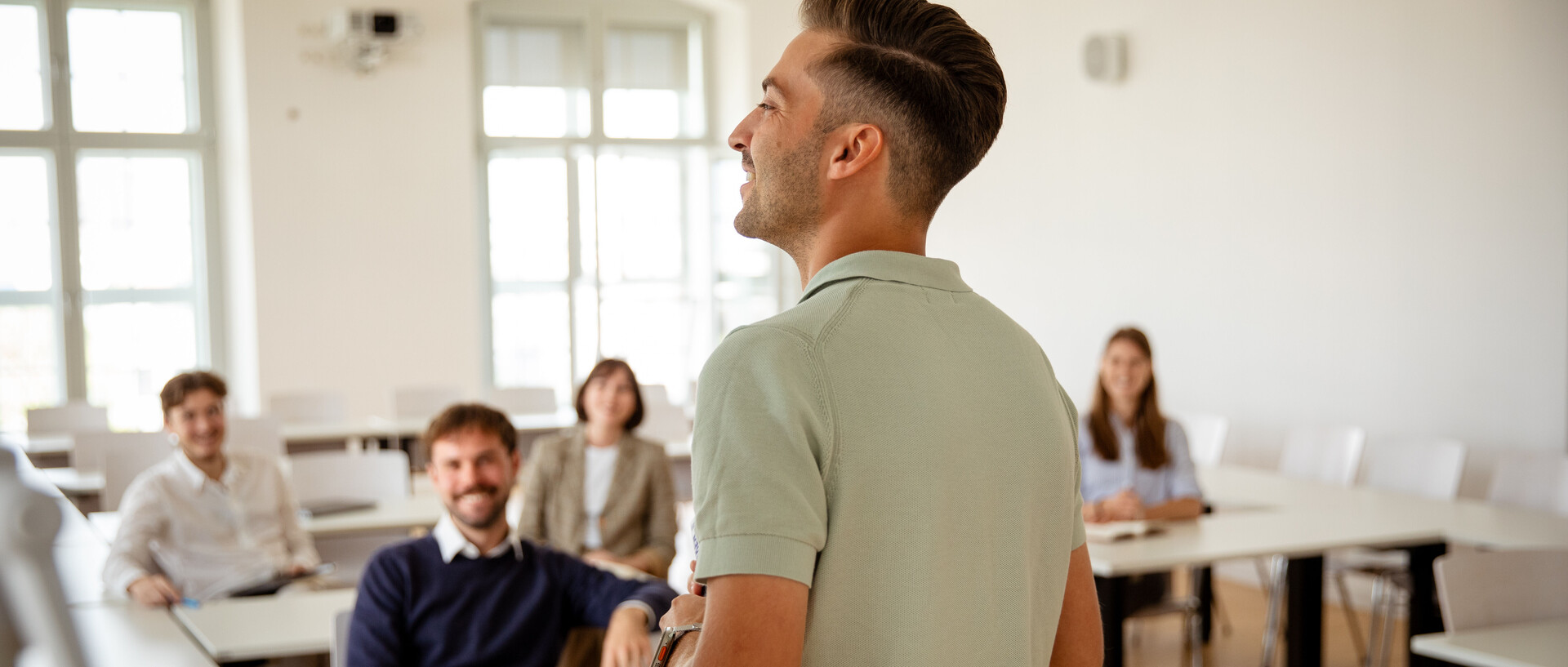 Eine Person in einem Poloshirt steht vorne in einem Semianrraum und redet. Fünf weitere Personen sitzen im Raum und sehen die Person an.