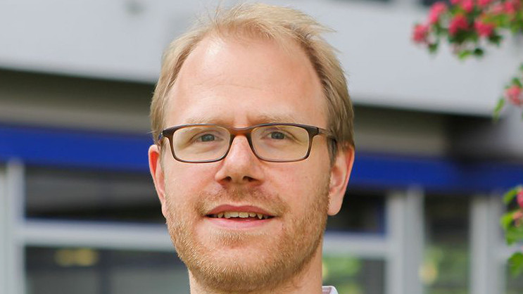 Professor Jochen Gebauer lächelt in die Kamera. Im Hintergrund ist das Gebäude A5.