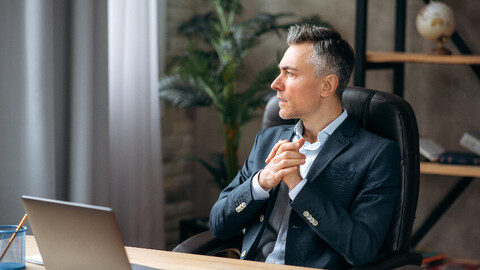 Eine Person in schicker Kleidung sitzt an einem Schreibtisch in einem Büro und blickt nach rechts. Vor ihm steht ein aufgeklappter Laptop.
