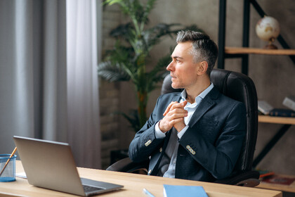 Eine Person in schicker Kleidung sitzt an einem Schreibtisch in einem Büro und blickt nach rechts. Vor ihm steht ein aufgeklappter Laptop.
