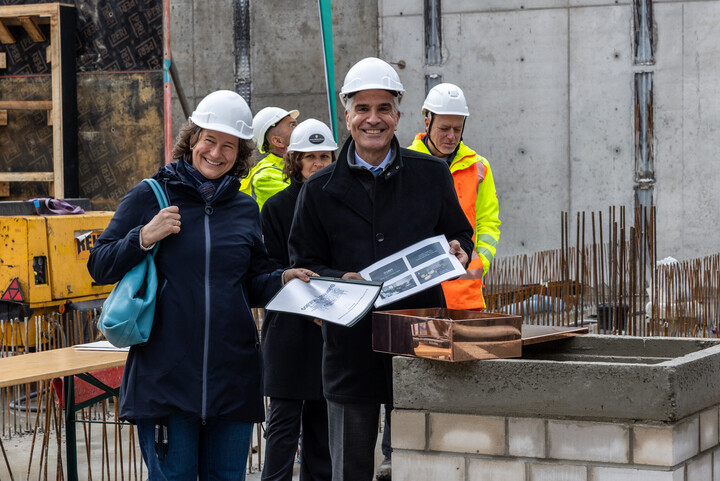 Dr. Katrin Schoppa-Bauer und Prof. Dr. Thomas Fetzer stehen auf der Baustelle, tragen Bauhelme und lachen in die Kamera. Prof. Fetzer hält Unterlagen in den Händen. Im Hintergrund stehen weitere Menschen mit Helmen und leuchtender Kleidung.