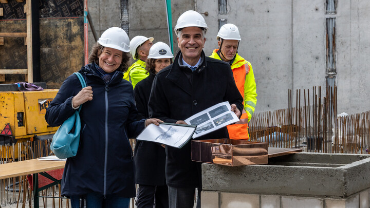 Dr. Katrin Schoppa-Bauer und Prof. Dr. Thomas Fetzer stehen auf der Baustelle, tragen Bauhelme und lachen in die Kamera. Prof. Fetzer hält Unterlagen in den Händen. Im Hintergrund stehen weitere Menschen mit Helmen und leuchtender Kleidung.