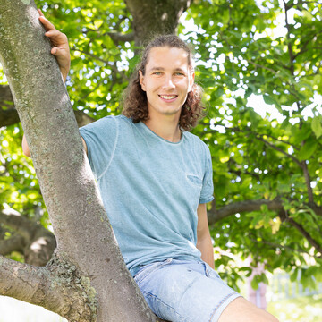  Luca Knörzer. Er sitzt lächelnd auf einem Ast. Er trägt ein hellblaues T-Shirt und seine Haare zur Hälfte hochgesteckt.