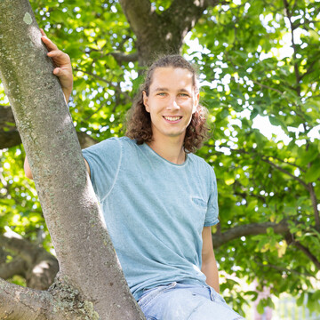  Luca Knörzer. Er sitzt lächelnd auf einem Ast. Er trägt ein hellblaues T-Shirt und seine Haare zur Hälfte hochgesteckt.