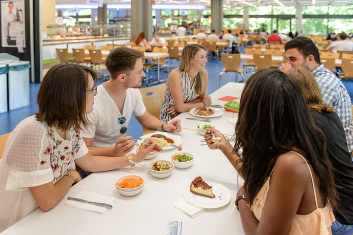 Eine Gruppe von Studierenden sitzt in der Mensa an einem Tisch. Sie reden und lachen. Auf dem Tisch stehen Teller mit Salat, Mittagessen und Kuchen. 