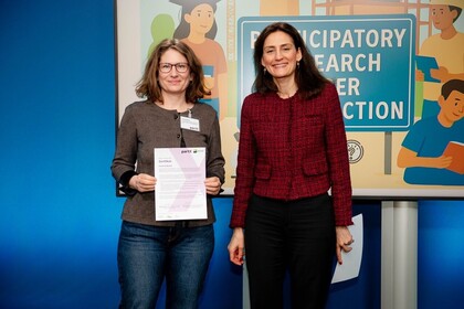 Zwei Frauen stehen nebeneinander vor einer Ausstellungswand zum Thema partizipative Forschung. Links steht Corinna Braun, Mitarbeiterin des ZLBI, sie hält ein Zertifikat in den Händen und trägt eine Brille sowie ein Namensschild. Rechts steht Dr. Effrosyni Chelioti, Abteilungsleiterin für Strategien und Grundsatzfragen im Bundesministerium, mit rotem Jackett. Beide blicken lächelnd in die Kamera.