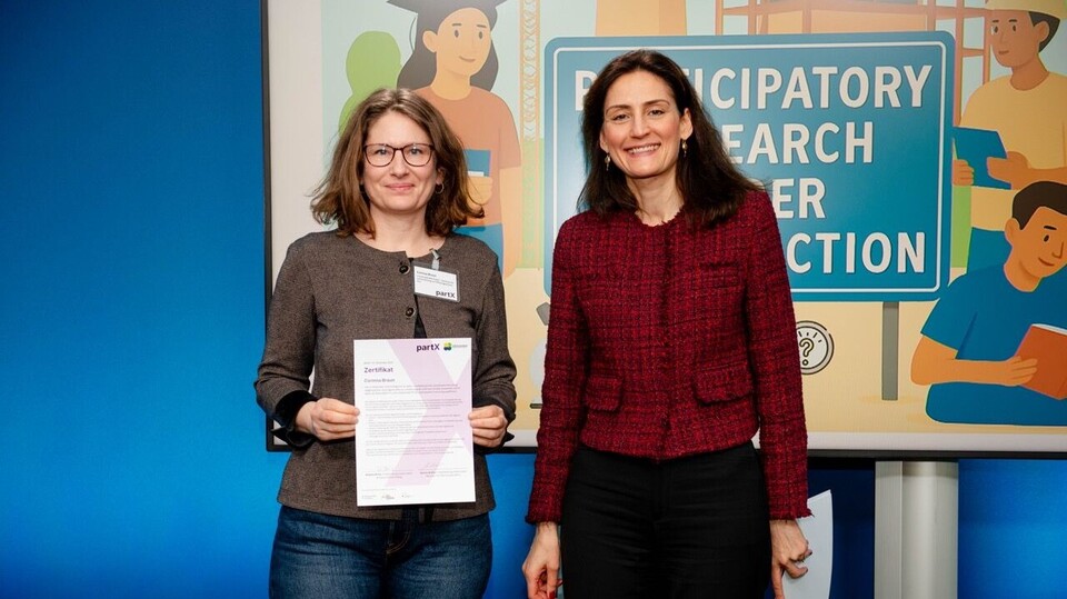 Zwei Frauen stehen nebeneinander vor einer Ausstellungswand zum Thema partizipative Forschung. Links steht Corinna Braun, Mitarbeiterin des ZLBI, sie hält ein Zertifikat in den Händen und trägt eine Brille sowie ein Namensschild. Rechts steht Dr. Effrosyni Chelioti, Abteilungsleiterin für Strategien und Grundsatzfragen im Bundesministerium, mit rotem Jackett. Beide blicken lächelnd in die Kamera.