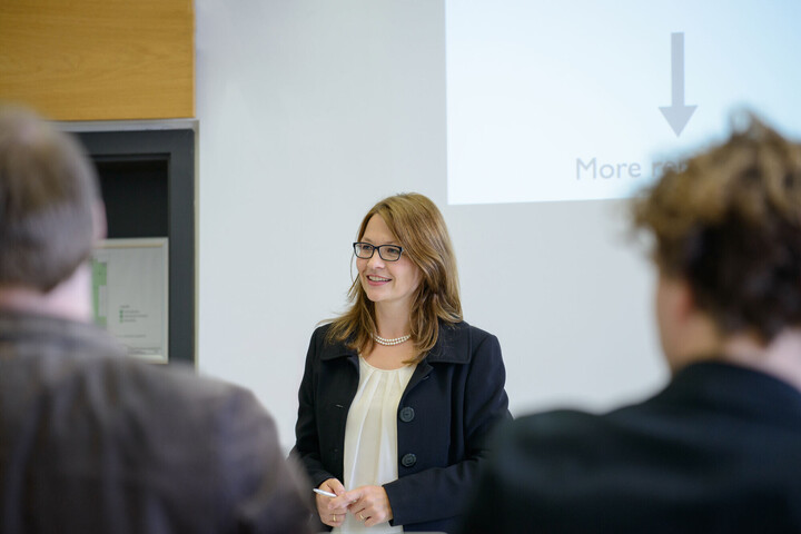 A woman giving a lecture. She has glasses and is wearing a black suit with a white blouse.