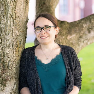  Veronique Embach. Sie lehnt lächelnd an einem Baum. Ihre Haare sind hochgesteckt, sie trägt eine Brille und einen dunklen Cardigan über dem türkisfarbenen Oberteil.