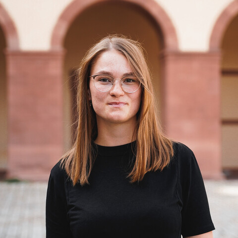 Eine Person trägt ein schwarzes T-Shirt und steht vor einem Schlossbogen der Uni Mannheim. Die Person heißt Johanna Mehltretter.