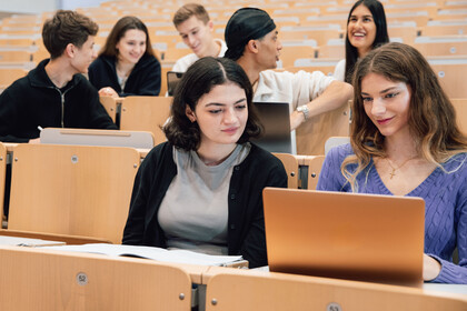 Studierende sitzen in einem Hörsaal. Sie unterhalten sich und schauen auf Laptops.